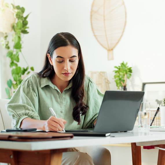 A woman sitting at her desk, writing and using her laptop.
